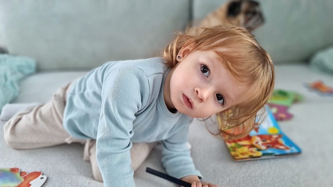 Adorable child kneels on a sofa, coloring with a pencil surrounded by toys in a cozy indoor setting.