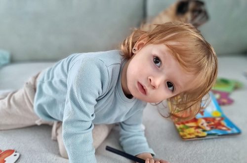 Adorable child kneels on a sofa, coloring with a pencil surrounded by toys in a cozy indoor setting.
