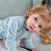 Adorable child kneels on a sofa, coloring with a pencil surrounded by toys in a cozy indoor setting.